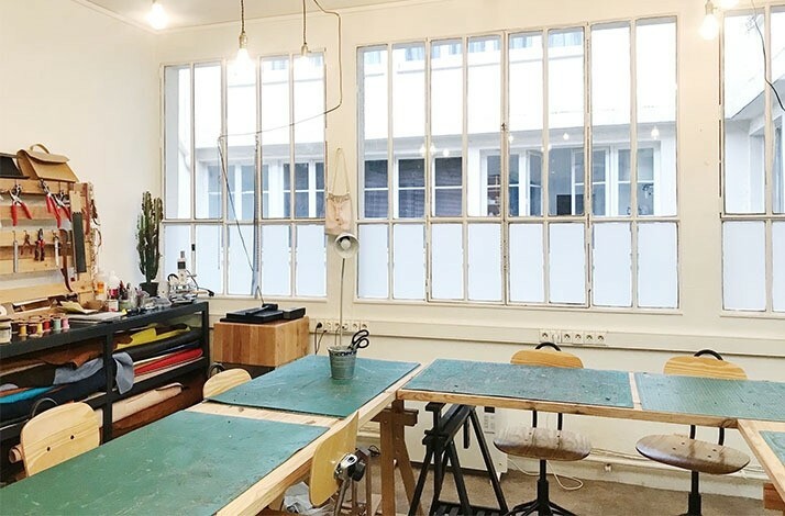 The interior of the Atelier La Patine in Paris with desks and chairs, shelves with leatherworking materials and a large window.