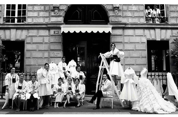 Black-and-white photo of a model posing in a Dior haut-couture and a team of tailors at the main entrance to the Maison Dior in Paris.